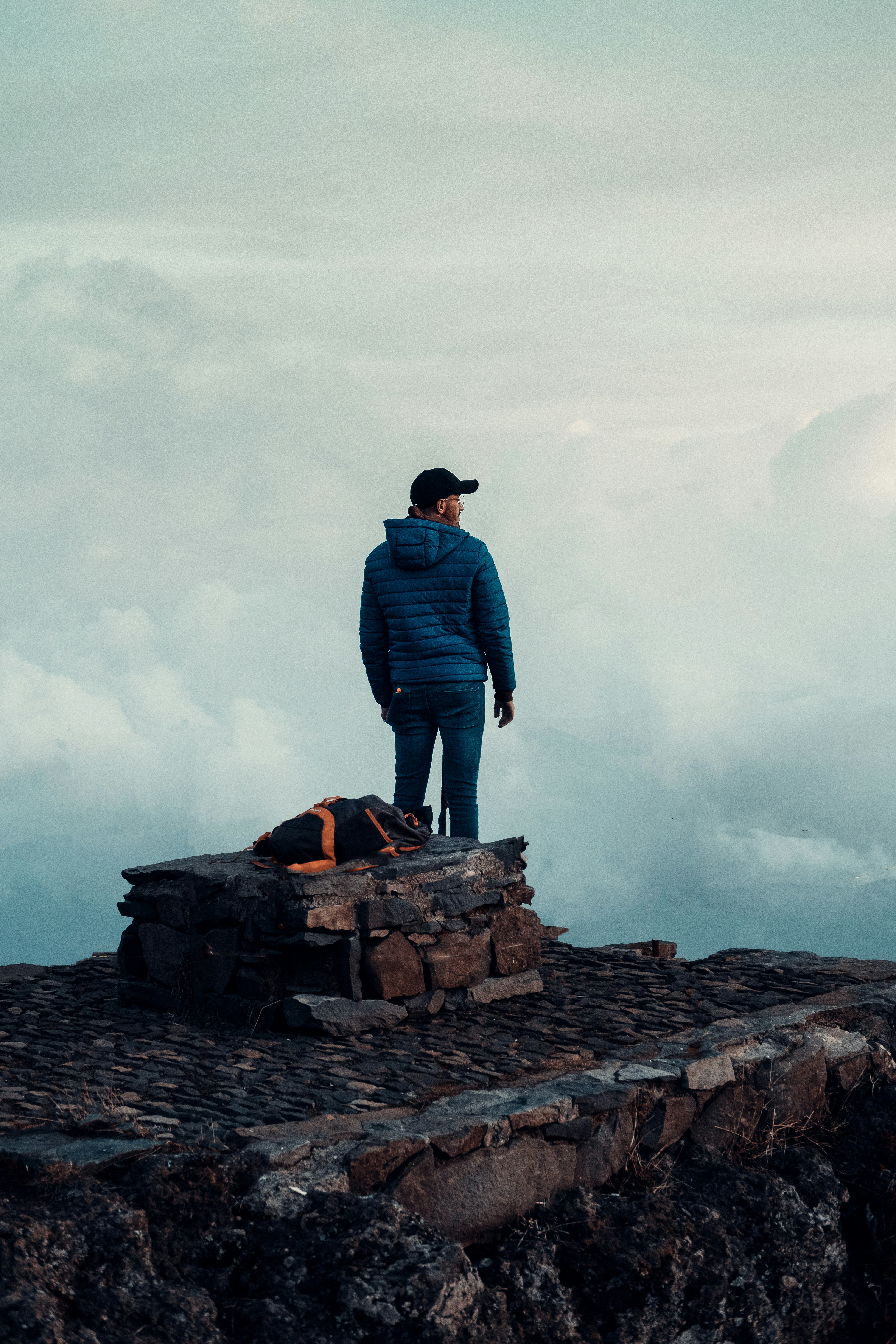 man in blue jacket standing on rock formation looking at white clouds during daytime